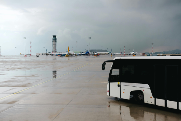 Airport tarmac with parked aircraft, control tower, and a ground transport bus under overcast skies.