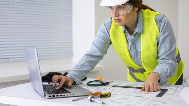 Engineer in a hard hat and safety vest reviewing blueprints on a laptop at a workstation.